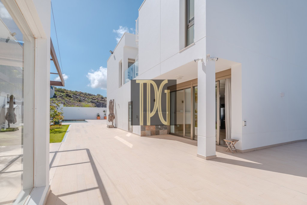 White modern house with a large glass-door patio, pool in the background, under a bright blue sky.
