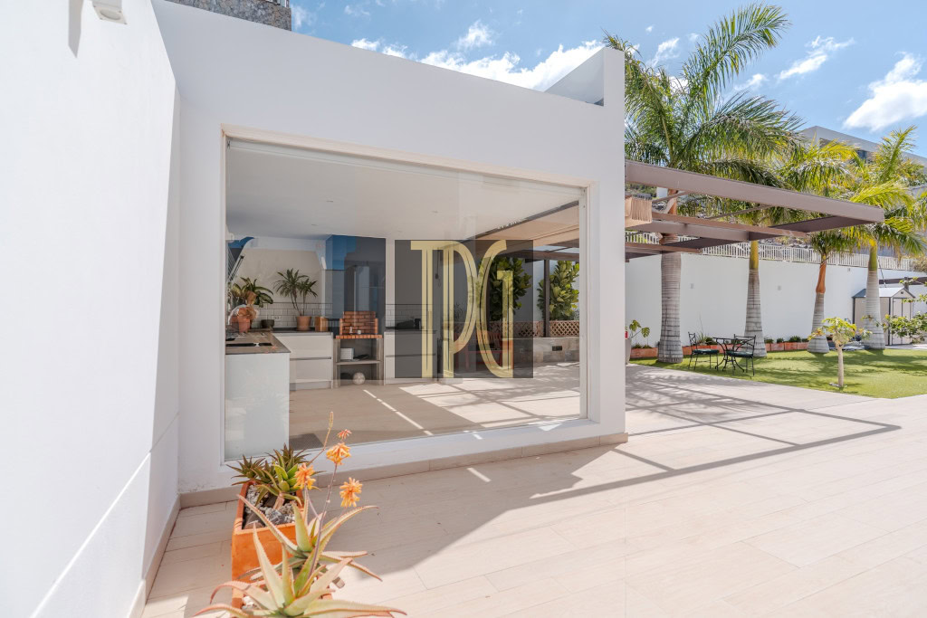 White modern house with a large glass door opening to an outdoor kitchen and patio, sunny day with palm trees in the yard