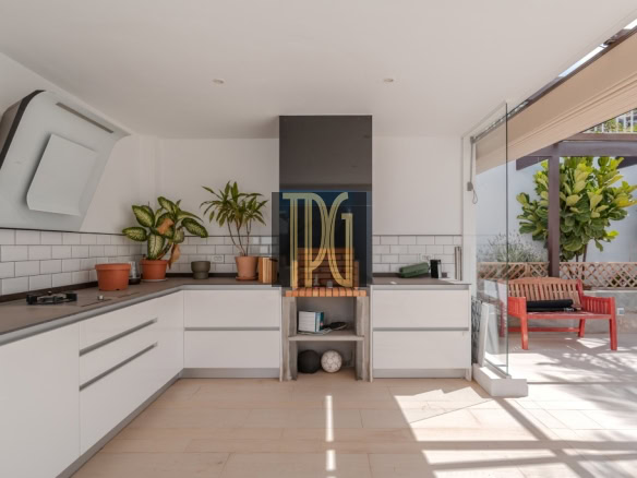 Modern white kitchen with lower cabinets, a central grill area, and potted plants on the counter, opening to a sunny outdoor seating area.