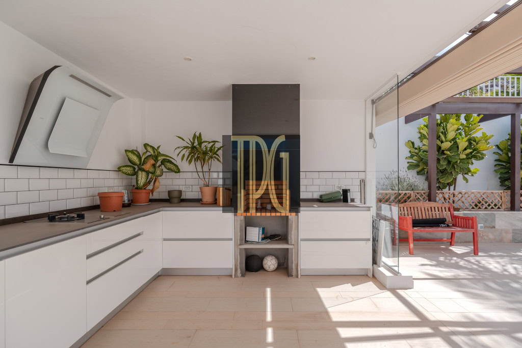 Modern white kitchen with lower cabinets, a central grill area, and potted plants on the counter, opening to a sunny outdoor seating area.