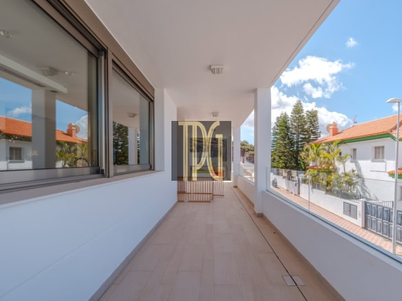 Long outdoor balcony with beige tile floor, white walls, and glass railing overlooking a sunny street with white houses and orange-tiled roofs.