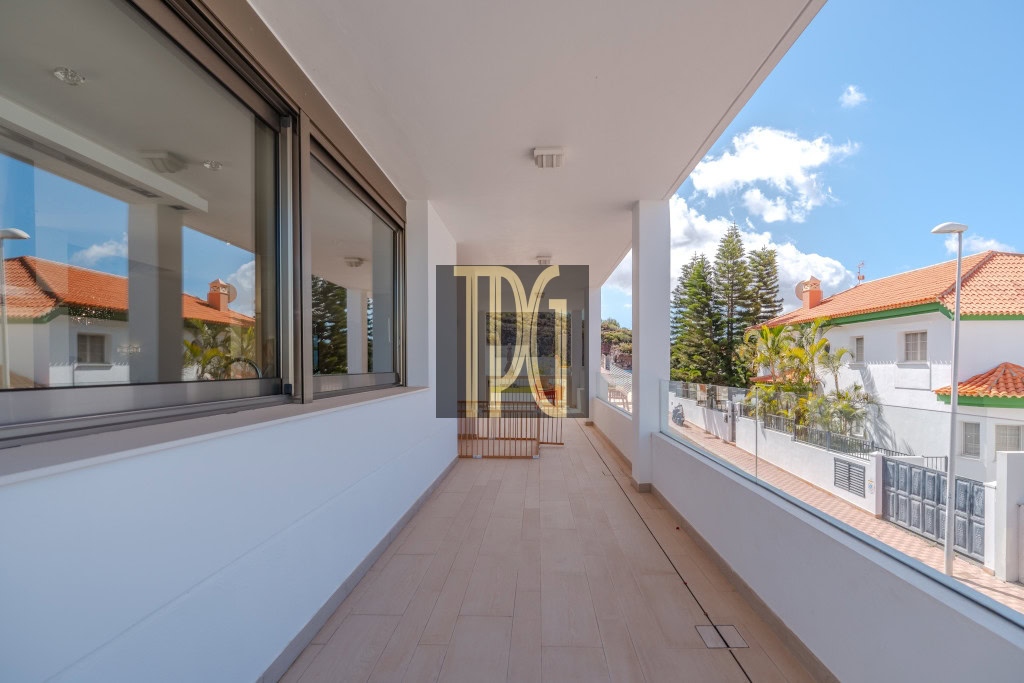 Long outdoor balcony with beige tile floor, white walls, and glass railing overlooking a sunny street with white houses and orange-tiled roofs.