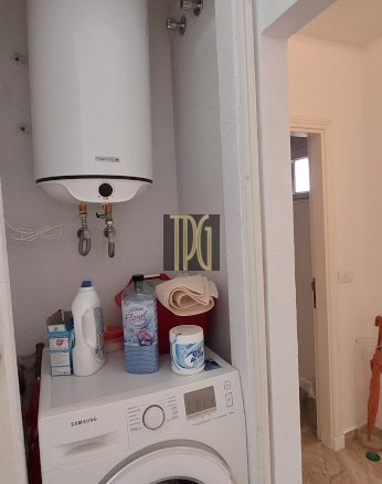 Tall white water heater mounted in a small closet above a front-loading Samsung washing machine; detergent bottles and a towel on the washer.