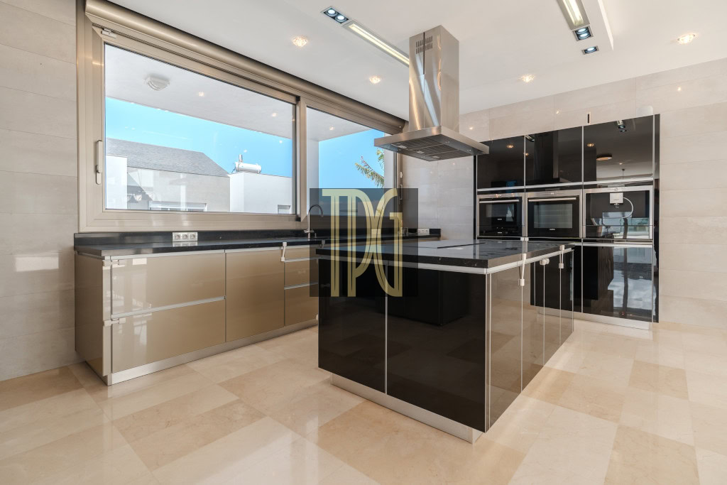 Modern kitchen with a black glossy island, stainless steel range hood, and built-in ovens along a wall of matching cabinets.