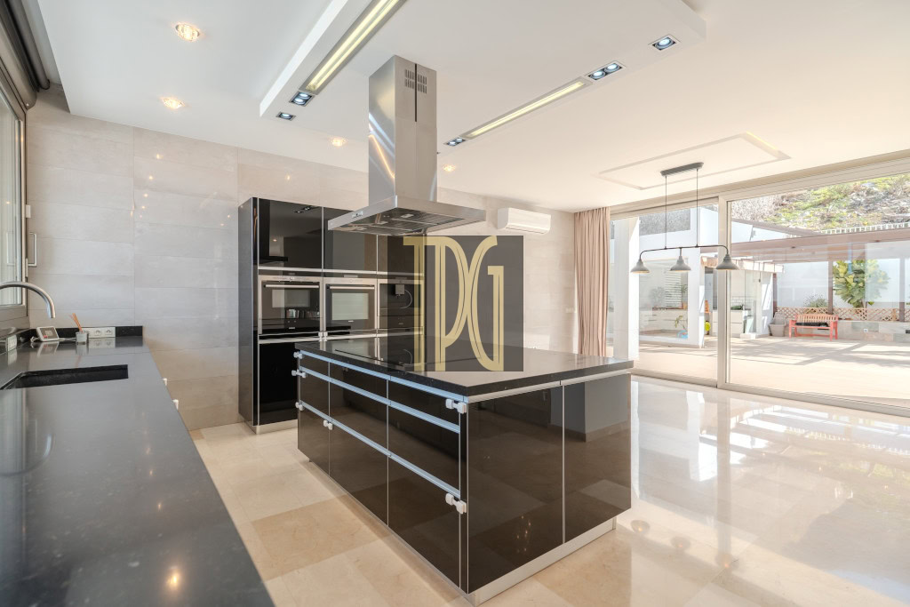 Modern kitchen with a glossy black island, stainless range hood, and built-in ovens; large glass doors to outdoor patio nearby.