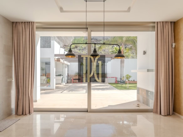 Modern living room with large glass sliding doors opening to a sunny backyard patio, flanked by beige curtains and a polished floor, with three pendant lights overhead.