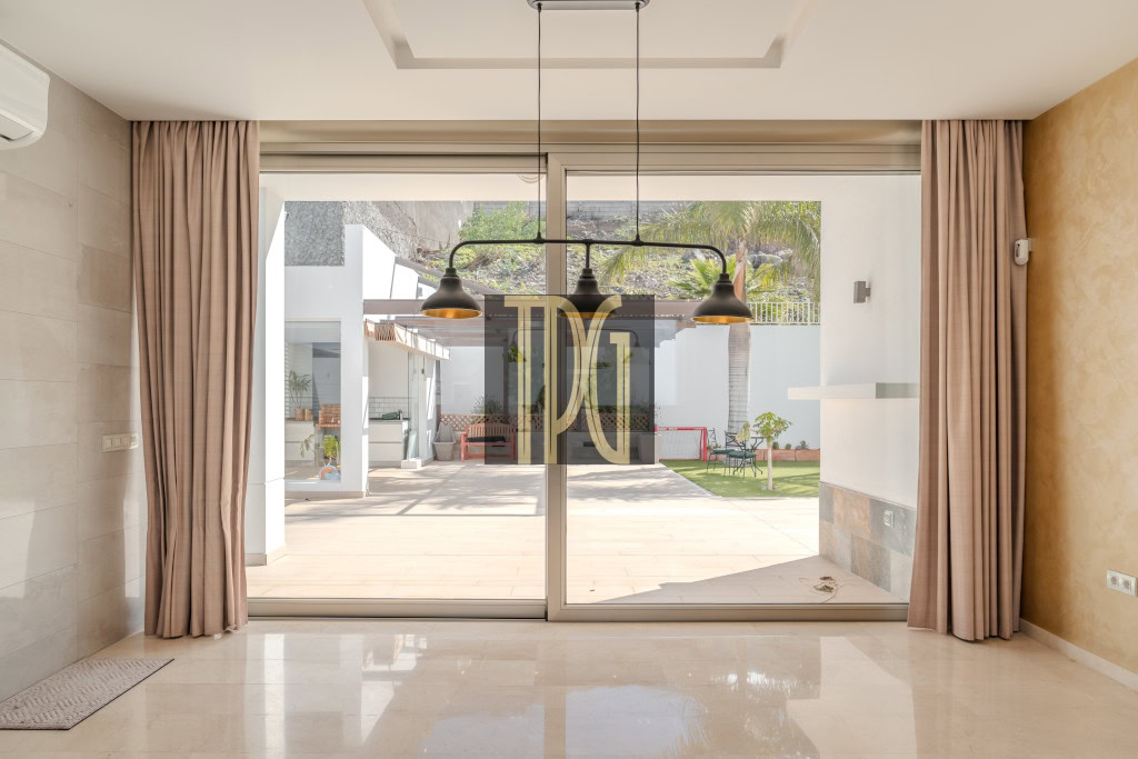 Modern living room with large glass sliding doors opening to a sunny backyard patio, flanked by beige curtains and a polished floor, with three pendant lights overhead.