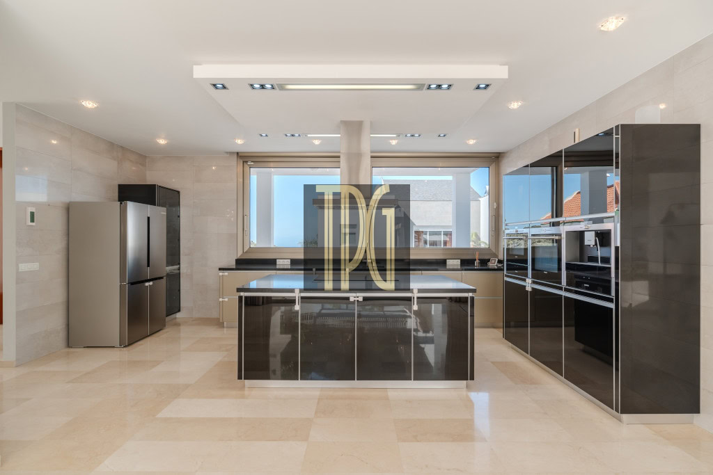 Modern, spacious kitchen with a large central island, glossy black cabinets, and stainless steel appliances including a tall fridge and double ovens, facing a wide window.