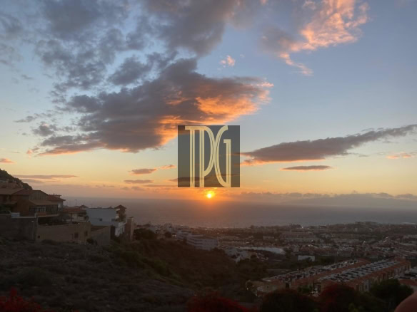 Sunset over a coastal hillside town with orange sun near the horizon and a calm sea in the distance.