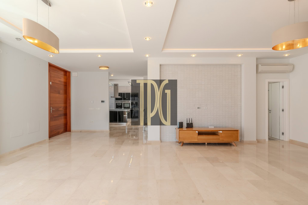 Empty modern living room with beige marble floor, wooden door on the left, and a low wooden TV console against a tiled wall in the center.