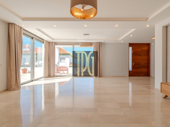 Bright empty living room with marble floor, beige curtains, and glass sliding doors opening to a sunlit patio outside.