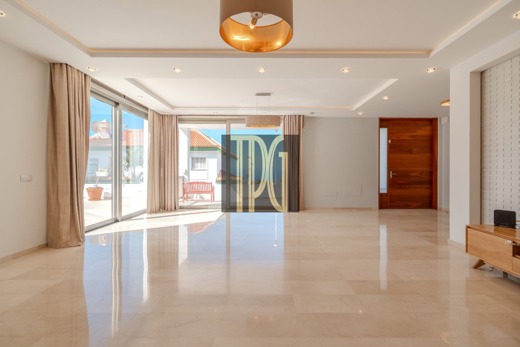 Bright empty living room with marble floor, beige curtains, and glass sliding doors opening to a sunlit patio outside.