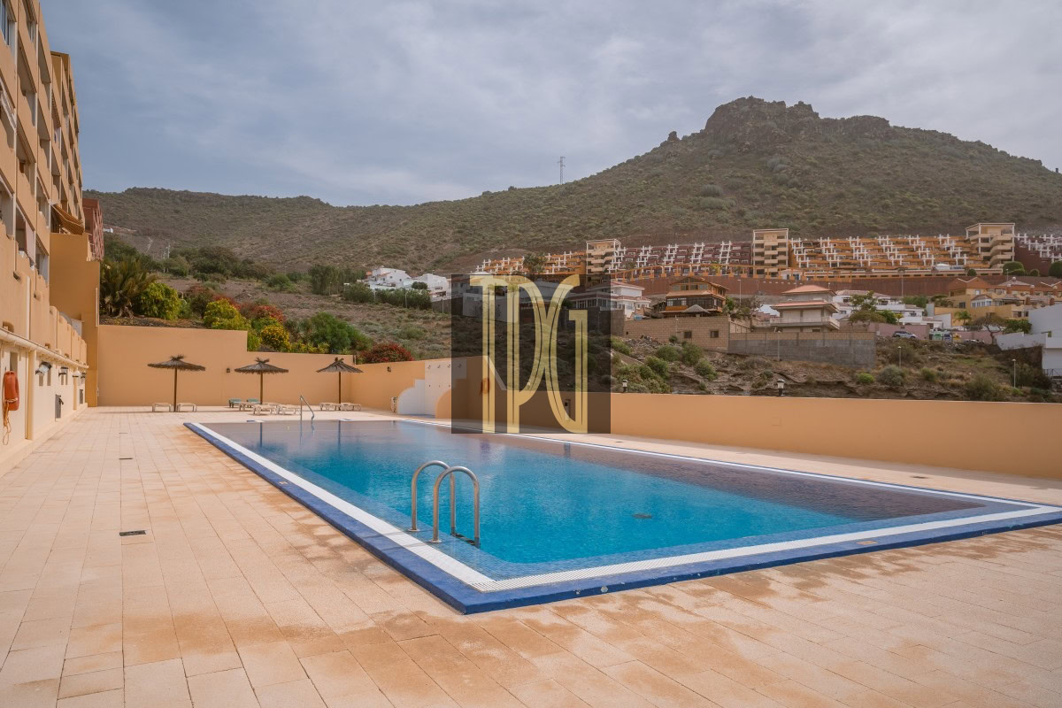 Rectangular blue pool in a resort courtyard with tan tile deck and beige walls, mountains in the background