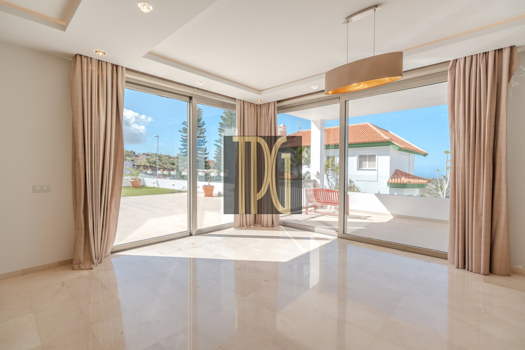 Sunlit living room with large glass sliding doors opening to a patio, beige curtains, and a polished tile floor.