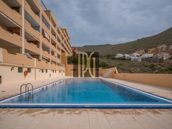 Rectangular blue swimming pool framed by beige apartment buildings on the left and a hillside in the distance under a clear sky.