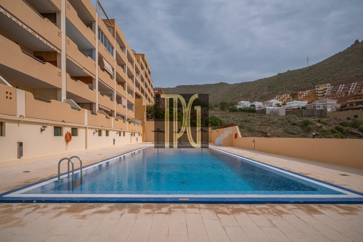 Rectangular blue swimming pool framed by beige apartment buildings on the left and a hillside in the distance under a clear sky.