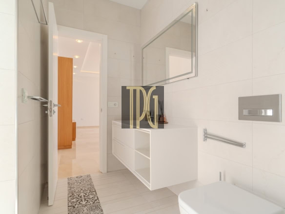 Bright modern bathroom with a white vanity, large mirror, and wall-mounted flush plate; door open to a light hallway, patterned floor tile strip nearby.