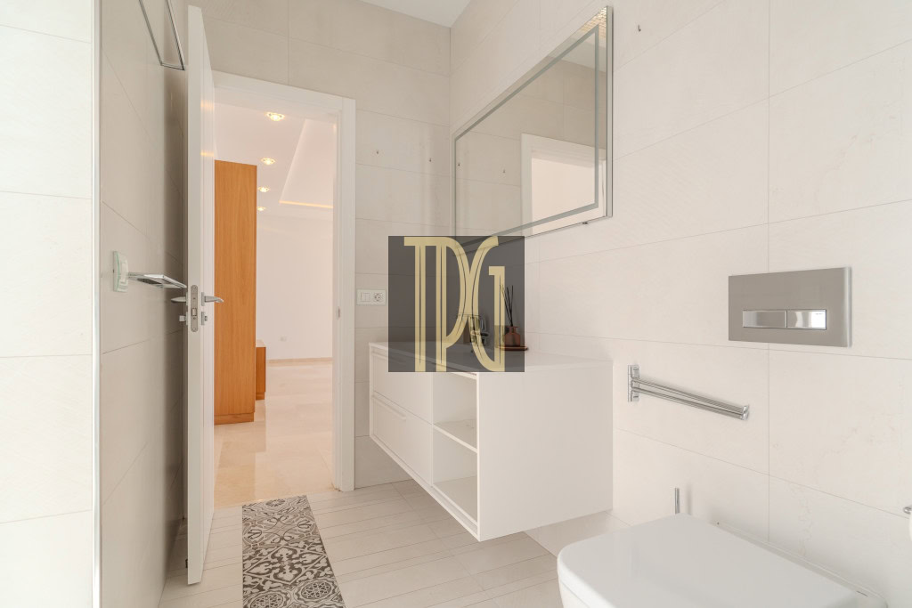 Bright modern bathroom with a white vanity, large mirror, and wall-mounted flush plate; door open to a light hallway, patterned floor tile strip nearby.