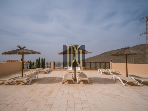 Sun loungers on a tiled terrace with thatched umbrellas and a railing overlooking hills and clear sky.
