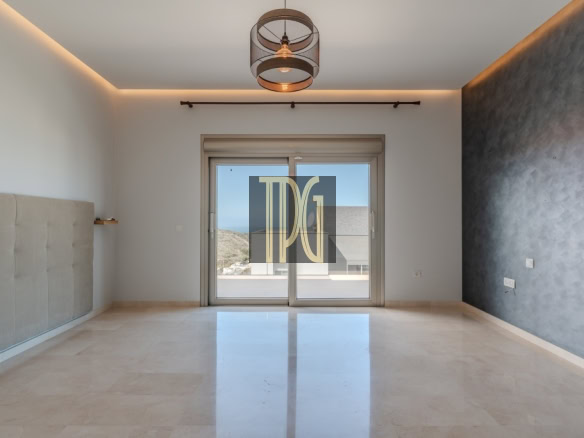 Empty modern living room with glossy beige tile floor, a padded headboard on the left wall, and a sliding glass door opening to a balcony; warm ceiling lights and a metal pendant chandelier illuminate the space.