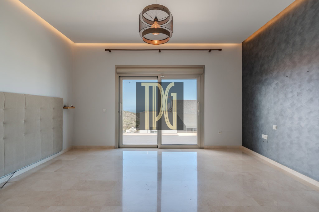Empty modern living room with glossy beige tile floor, a padded headboard on the left wall, and a sliding glass door opening to a balcony; warm ceiling lights and a metal pendant chandelier illuminate the space.