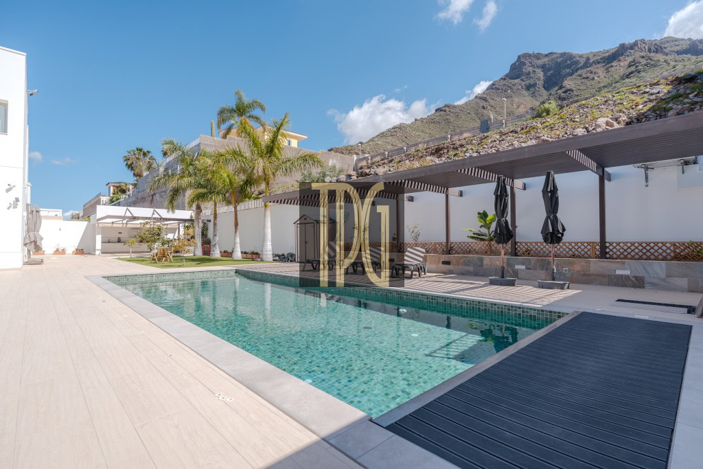 Outdoor pool area at a sunny resort with palm trees, lounge chairs, and shaded seating under a pergola.