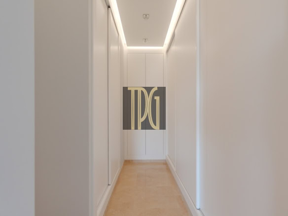 Narrow white hallway with recessed ceiling lights and a beige stone floor, ending at a white cabinet.