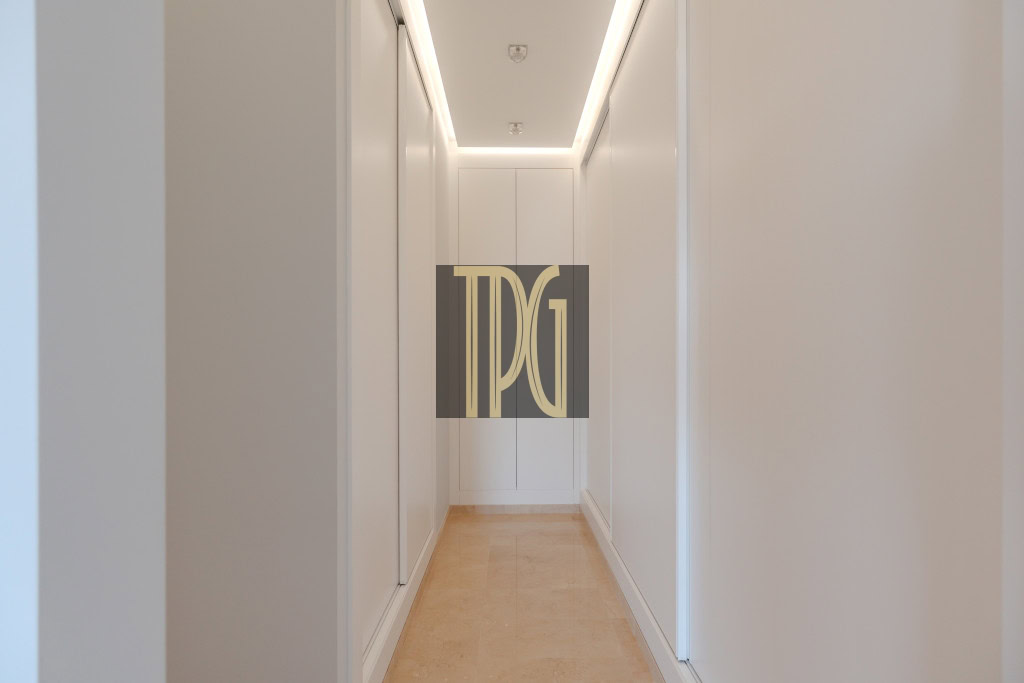 Narrow white hallway with recessed ceiling lights and a beige stone floor, ending at a white cabinet.