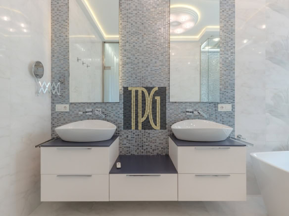 Twin white vessel sinks on a dark countertop with a mosaic tile backsplash and a large monogram panel between mirrors in a modern bathroom, with a tub visible to the right.