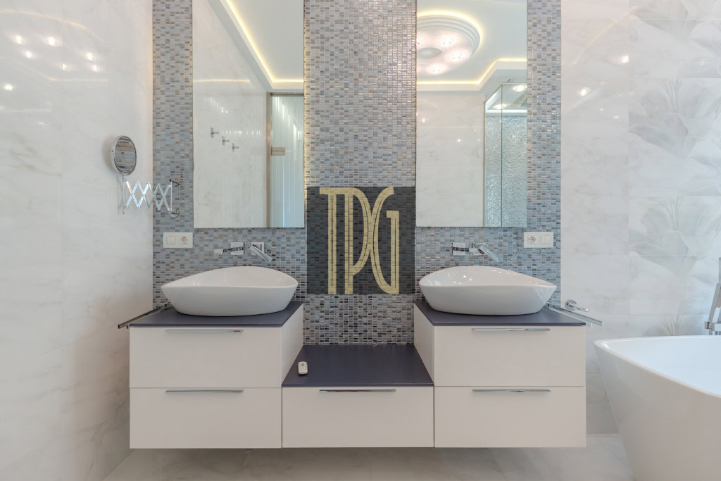 Twin white vessel sinks on a dark countertop with a mosaic tile backsplash and a large monogram panel between mirrors in a modern bathroom, with a tub visible to the right.