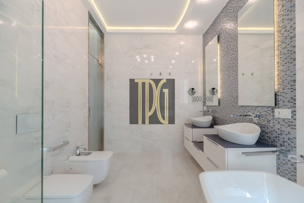Modern white bathroom with a glass shower, bidet and toilet on the left, and a floating vanity with two vessel sinks along a mosaic-tiled wall on the right.