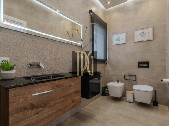 Modern bathroom with a wooden vanity, black countertop, wall faucet, and a large backlit mirror; plant on counter nearby.
