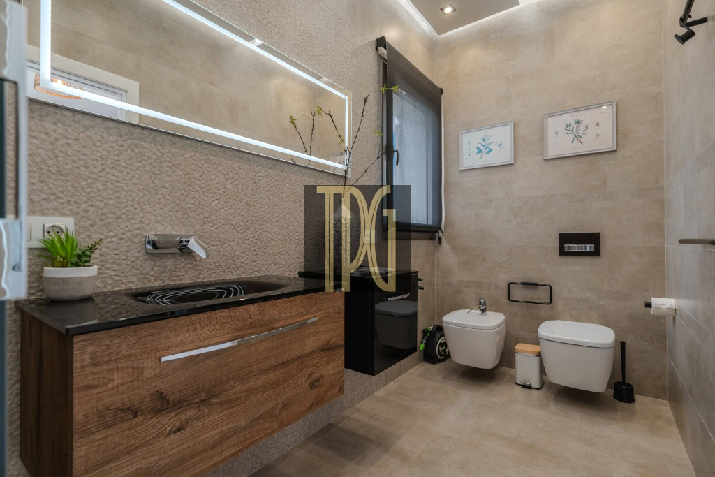 Modern bathroom with a wooden vanity, black countertop, wall faucet, and a large backlit mirror; plant on counter nearby.