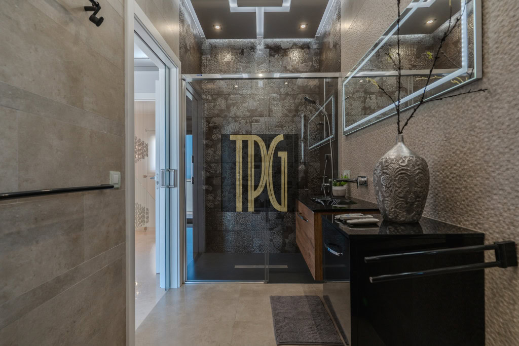 Modern bathroom with glass shower door featuring a large gold TDG logo; black countertop and decorative vase nearby
