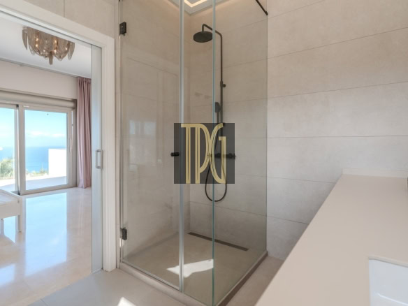 Modern bathroom with a glass-enclosed shower and white countertop, opening to a bright room with an ocean view.