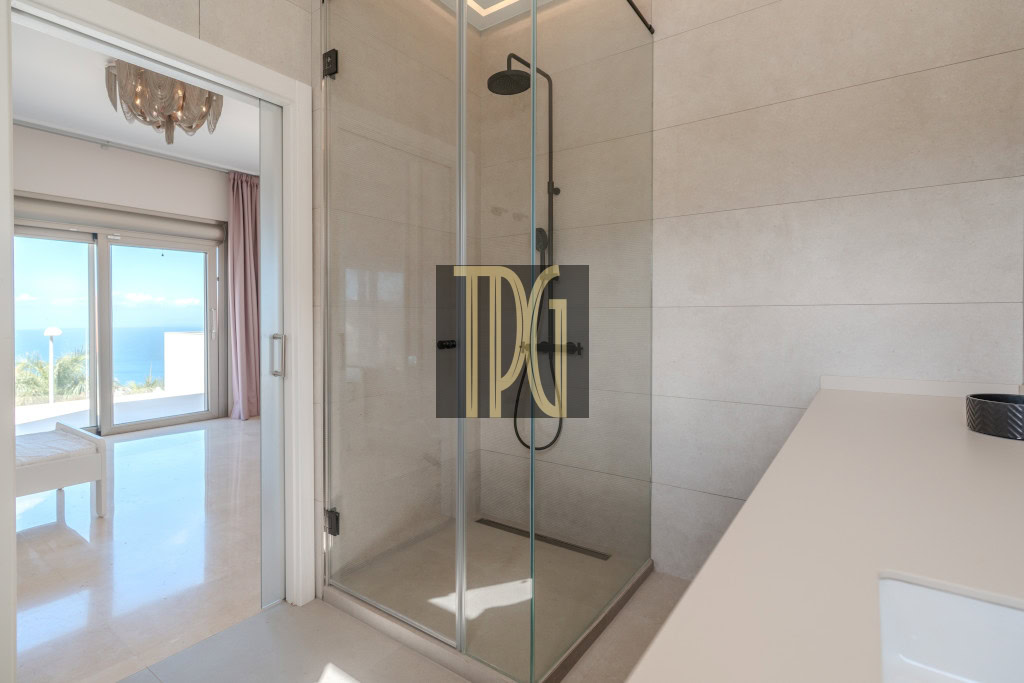 Modern bathroom with a glass-enclosed shower and white countertop, opening to a bright room with an ocean view.