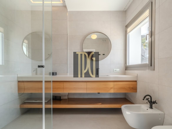 Modern bathroom with a round mirror above a floating wooden vanity, glass shower partition, and a white bidet by a window with natural light.
