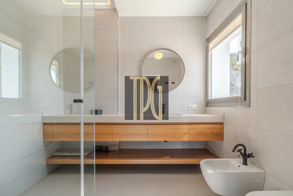 Modern bathroom with a round mirror above a floating wooden vanity, glass shower partition, and a white bidet by a window with natural light.