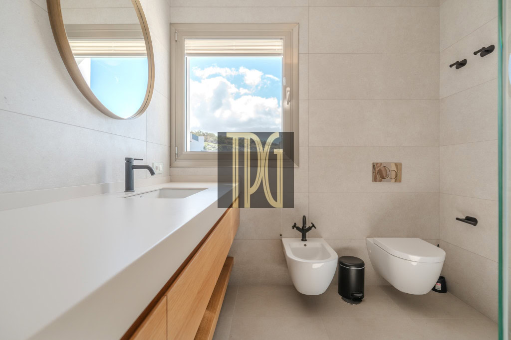Modern bathroom with white countertop sink, round mirror, window, and a bidet beside a wall-mounted toilet.