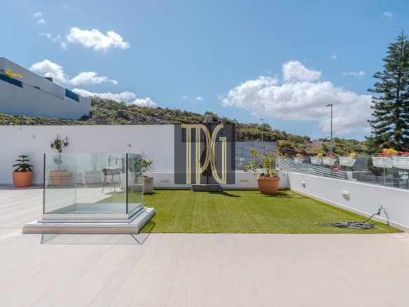 Sunlit rooftop terrace with artificial grass, glass railing, potted plants, and a clear blue sky.