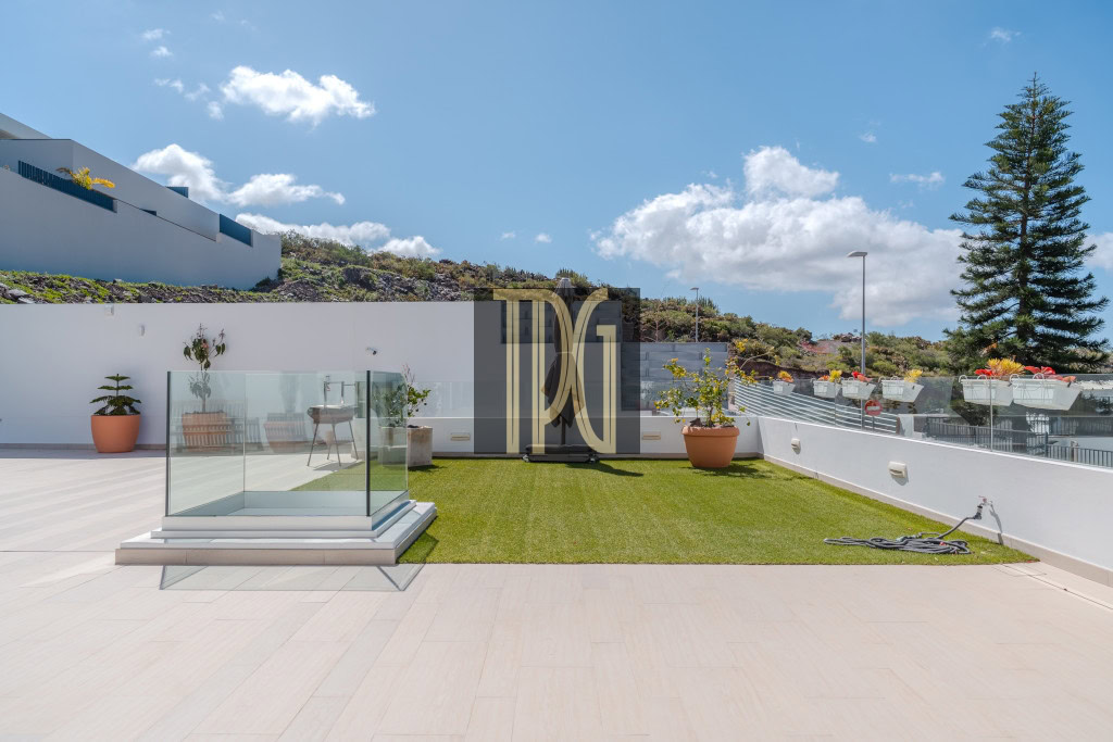 Sunlit rooftop terrace with artificial grass, glass railing, potted plants, and a clear blue sky.