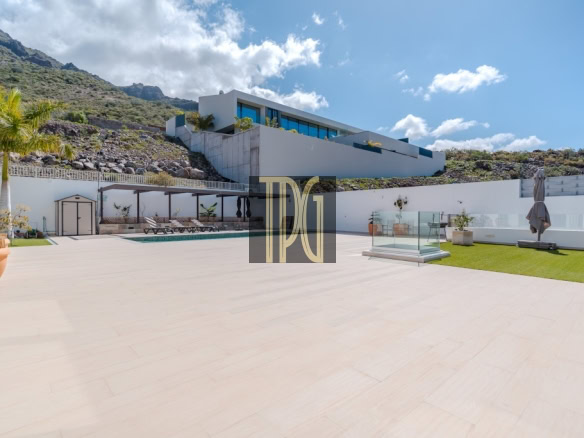 Modern villa terrace with a rectangular pool, lounge chairs, and glass railing against a hillside and blue sky.