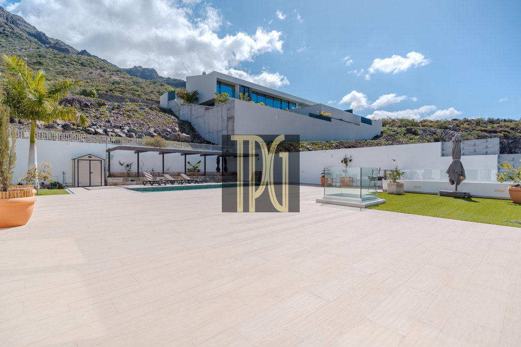 Modern villa terrace with a rectangular pool, lounge chairs, and glass railing against a hillside and blue sky.