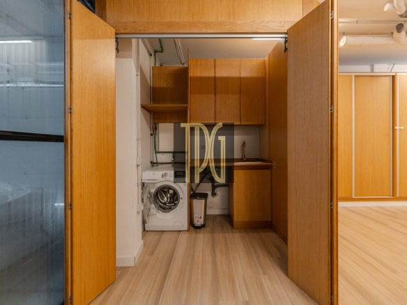 Laundry nook behind open wooden doors: washing machine, sink, and upper cabinets on a wood floor.