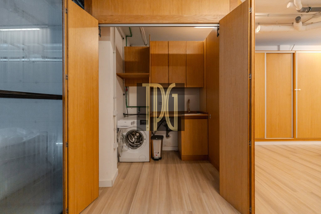 Laundry nook behind open wooden doors: washing machine, sink, and upper cabinets on a wood floor.