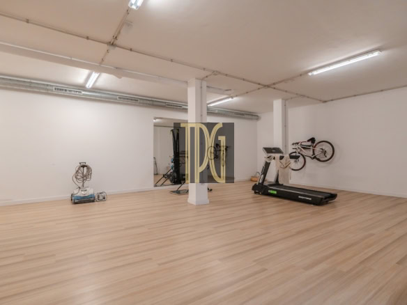 Spacious gym room with light wood floor, a treadmill, and bikes mounted on the right wall under bright ceiling lights, with a white column in the center.