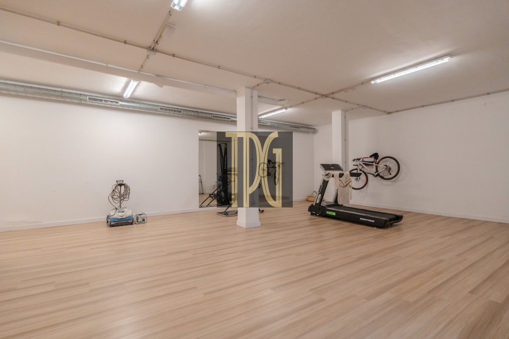 Spacious gym room with light wood floor, a treadmill, and bikes mounted on the right wall under bright ceiling lights, with a white column in the center.