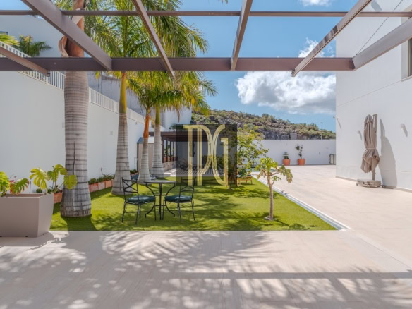 Open-air courtyard with palm trees, a black metal table and chairs on artificial grass, and white walls with a metal pergola overhead.