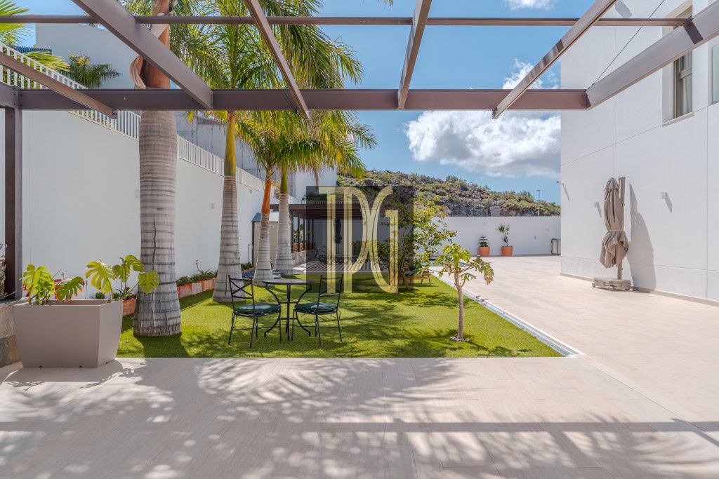 Open-air courtyard with palm trees, a black metal table and chairs on artificial grass, and white walls with a metal pergola overhead.