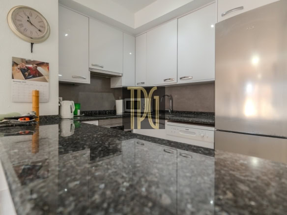 Modern white kitchen with glossy cabinets, dark granite counters, a stainless steel fridge and dishwasher, a wall clock and calendar on the wall, and a kettle on the counter.